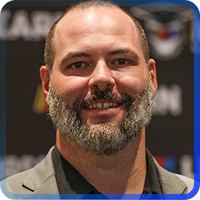 A headshot of a smiling. The photo is set against a plain beige background and framed by a thin, blue-to-gray gradient border.